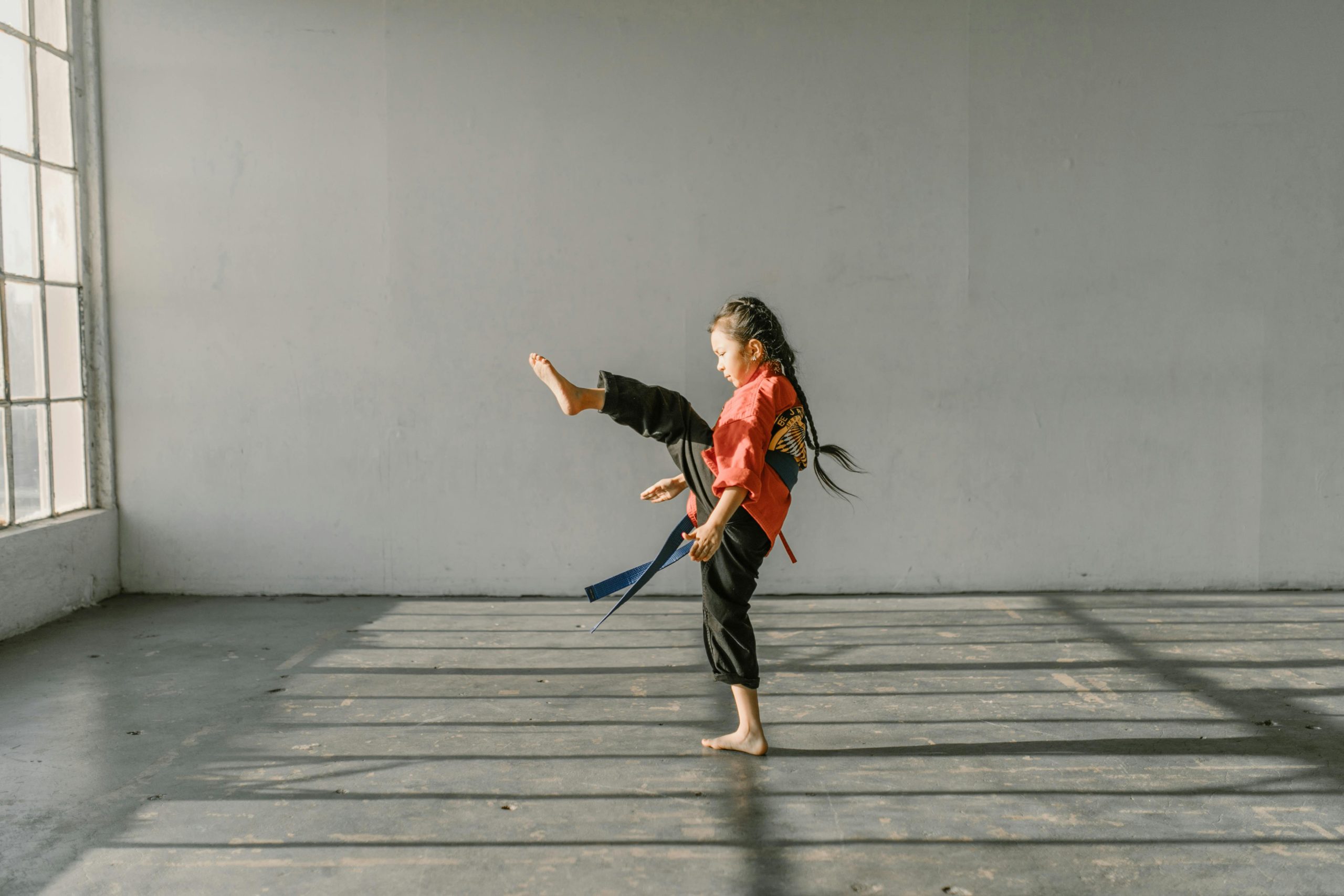 A young girl practicing a martial arts kick in a sunlit dojo indoor environment.